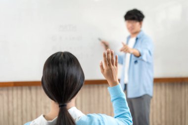 Beyaz tahtanın önünde duran erkek öğretmen öğrenciyi soru sormak için işaret ediyor ve genç öğrenciler sınıfta öğrenirken bir soruyu cevaplamak için ellerini havaya kaldırıyor.