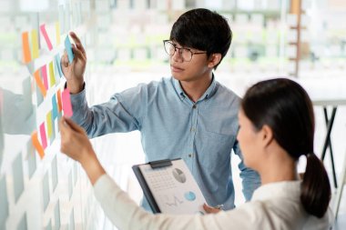 İki işletme yöneticisi, ofiste birlikte pazarlama planının planlanmasına yönelik yeni bir iş projesi hakkında fikir alışverişinde bulunmak için kilit noktayı duvara yazıp not alıyorlar..