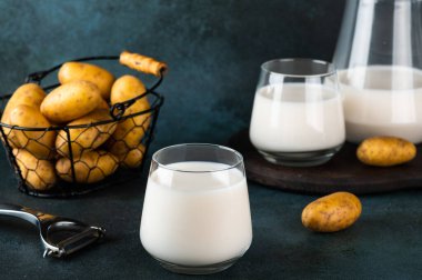 Vejetaryen patates sütünü bardağa, patatesi de koyu arkaplana döküyorum. Vejetaryen sütü. Bitki bazlı süt değiştirici ve laktozsuz. Sebze sütü.