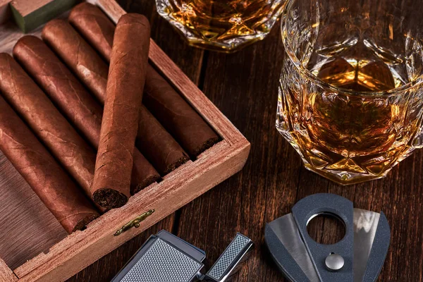 Box with cuban cigars, lighter and cutter on old wooden table top. Two glasses of whiskey or alcohol on the background.