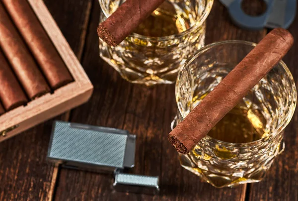 View of a wooden box with Cuban cigars, a lighter and a cutter. Still life with two glasses of whiskey on the table.