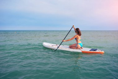 Mavi bikinili bir kadın güneşli bir günde denizde kürekle yemek masasında oturur.