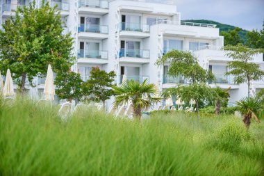 Birçok terası ve yeşil bahçesi olan beyaz bina oteli ön planda, tatil beldesi