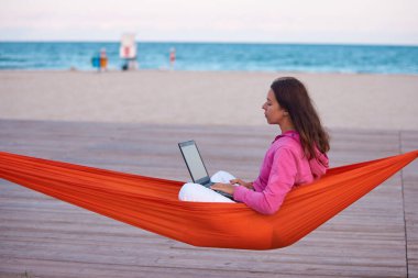 Young woman sitting on hammock and working remote at laptop computer on sea beach at sunset