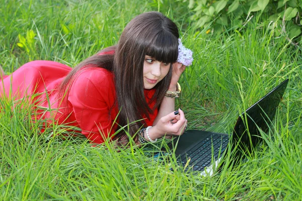 Το κορίτσι σε ένα κόκκινο φόρεμα με φορητό υπολογιστή που βρίσκεται στο γρασίδι — Φωτογραφία Αρχείου