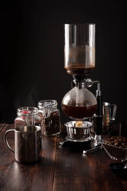 Vacuum coffee maker also known as vac pot, siphon or syphon coffee maker. Metallic cup and toasted coffee beans on rustic wooden table clipart