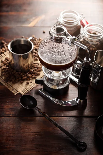 Vacuum coffee maker also known as vac pot, siphon or syphon coffee maker. Metallic cup and toasted coffee beans on rustic wooden table