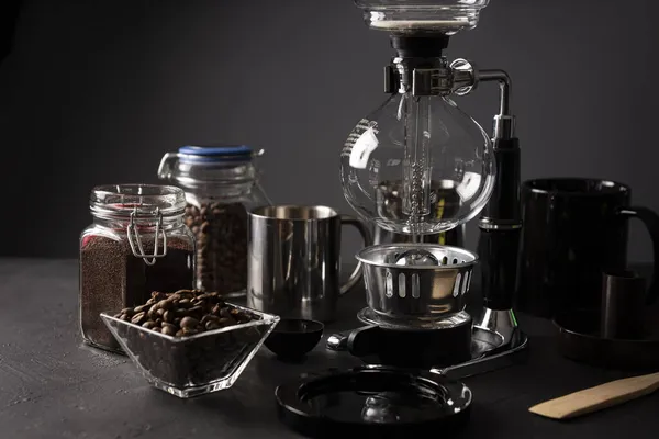 Vacuum coffee maker also known as vac pot, siphon or syphon coffee maker. Metallic cup and toasted coffee beans on rustic black stone table.