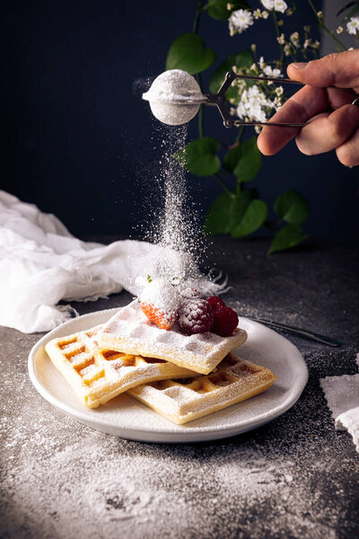 Human hand sprinkling powdered sugar on homemade waffles with strawberries and raspberries on white dish.