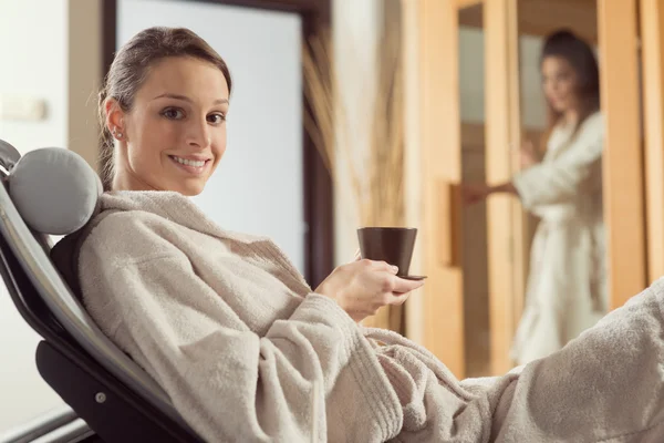 Mujer joven sonriendo y relajándose en el spa, sosteniendo una bebida caliente y saludable . — Foto de Stock