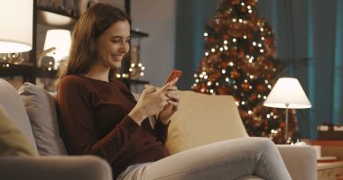 Oturma odasındaki kanepede oturan ve arka planda Noel ağacı olan akıllı telefonuyla sohbet eden mutlu genç kadın.