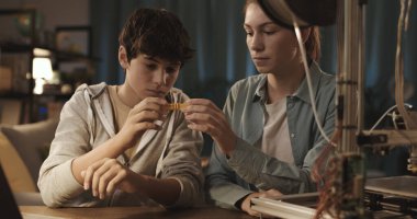 Masada oturan ve evde 3D baskı öğrenen öğrenciler plastik bir prototipi kontrol ediyorlar.