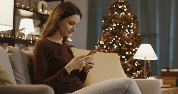 幸せな若いです女性座っているソファの上にリビングルームと彼女のスマートフォンとチャット 背景にクリスマスツリー — ストック写真
