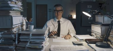 Gülümseyen bir ofis çalışanı, masa başında oturuyor ve tekrarlayan bir iş yapıyor, dosyaları damgalıyor.