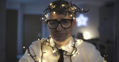 Hüzünlü iş adamı Noel günü çalışıyor, Noel ışıklarına sarılmış ve kameraya bakıyor.