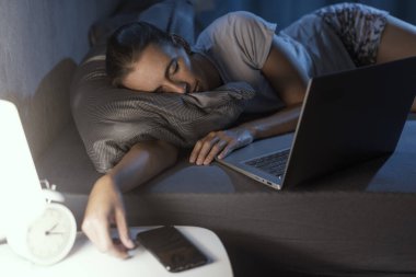 Yorgun bir kadın yatağında dizüstü bilgisayarının önünde uyuyakalıyor.