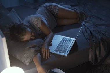 Yorgun bir kadın yatağında dizüstü bilgisayarının önünde uyuyakalıyor.