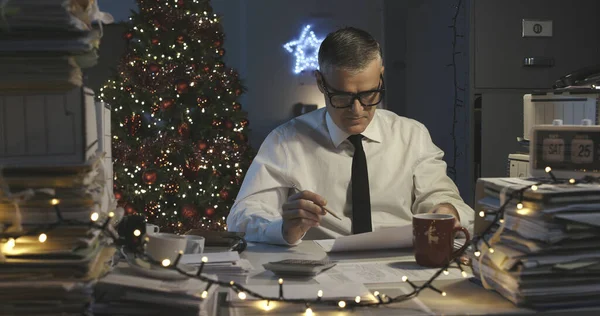 クリスマスの日に働く悲しいビジネスマン 彼は書類の山に囲まれています — ストック写真