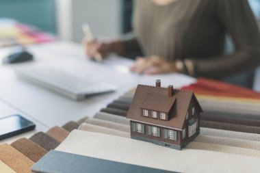 İç mimar masasında oturuyor ve çalışıyor, kumaş örnekleri ve model ev ön planda, seçici odaklı