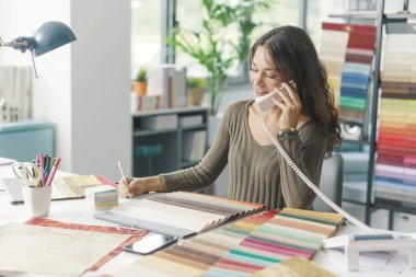 Genç yaratıcı dekoratör masada oturuyor ve çalışıyor. Bir telefon görüşmesi yapıyor ve notlar yazıyor.