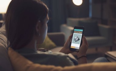 Evde dinlenen ve akıllı telefonuyla bağlantı kuran genç bir kadın bir alışveriş uygulamasında ikinci el eşyalar arıyor.