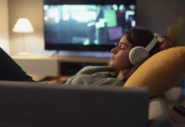 年轻女子躺在家里的沙发上 戴着无线耳机 听着音乐 — 图库照片