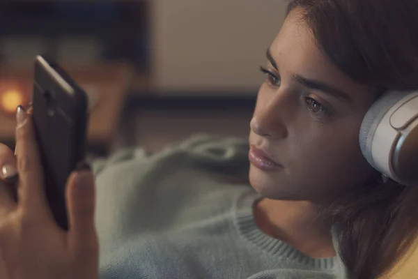 年轻的女人躺在沙发上休息 她戴着耳机 用智能手机连接着 — 图库照片