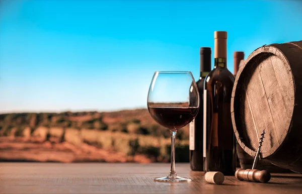 红酒酒杯 酒瓶和酒桶放在桌子上 葡萄园和蓝天放在背景上 — 图库照片