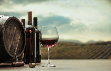 Mükemmel kırmızı şarap tatma deneyimi: şarap kadehi, şarap şişesi fıçısı masada ve üzüm bağı arka planda
