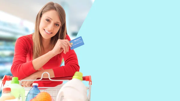 スーパーで食料品の買い物をしている笑顔の女性彼女はフルカートにもたれており クレジットカードを保持しています 空白のコピースペース — ストック写真