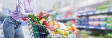 Kadın alışveriş arabasına yaslanıyor ve listeyi kontrol ediyor, market bayrağı.