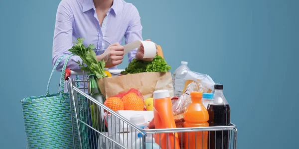 女性は完全なショッピングカートにもたれ 食料品の領収書をチェックします 費用と消費者主義の概念 — ストック写真