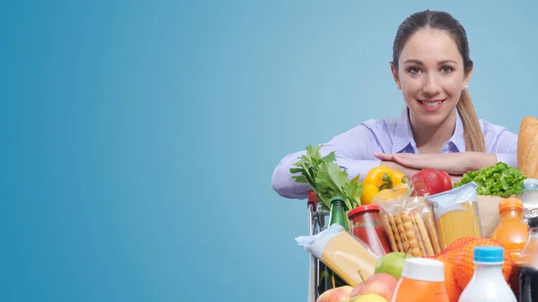 Glimlachende Jonge Vrouw Leunend Een Volle Winkelwagen Poserend Boodschappenconcept — Stockfoto