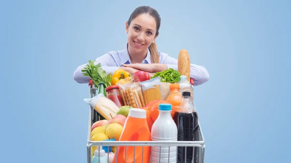 Lächelnde Junge Frau Die Sich Auf Einen Vollen Einkaufswagen Lehnt — Stockfoto