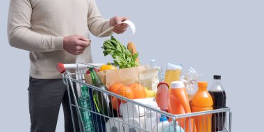 Adam alışveriş arabasının yanında duruyor ve market fişini kontrol ediyor.