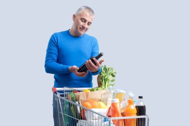Süpermarkette market alışverişi yapan bir adam ön planda tam teşekküllü bir alışveriş arabası ve bir şişe kırmızı şarap alıyor.