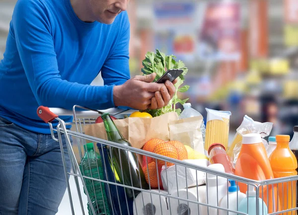 Homem Fazendo Compras Supermercado Ele Está Empurrando Carrinho Usando Smartphone — Fotografia de Stock