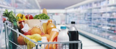 Yiyecek ve içeceklerle dolu alışveriş arabası ve arka planda süpermarket rafları, market alışverişi konsepti