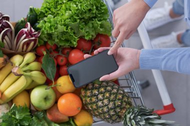 Market alışverişi yapan ve akıllı telefonunu kullanan bir kadın. Alışveriş arabası taze sebze ve meyvelerle dolu.