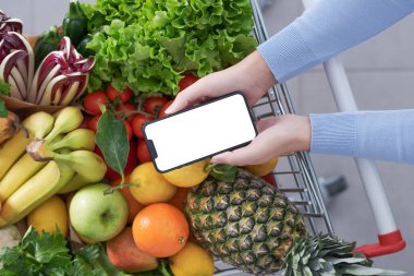 Market alışverişi yapan ve akıllı telefonunu kullanan bir kadın. Alışveriş arabası taze sebze ve meyvelerle dolu.