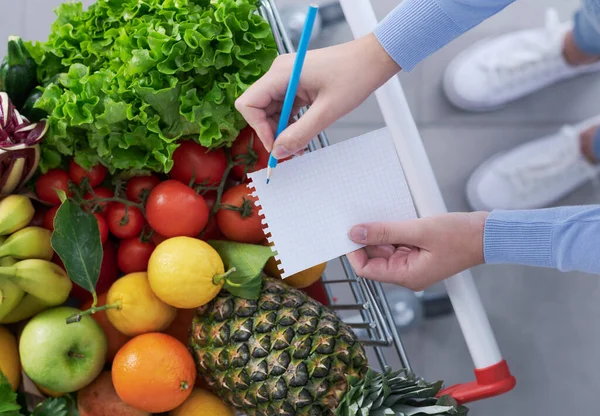 女人推着一辆满载新鲜蔬菜的购物车 查看购物单 健康食品和食品杂货购物的概念 — 图库照片