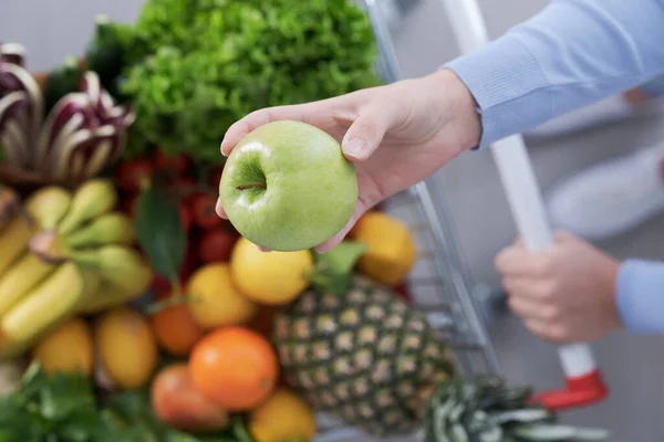 女人推着一辆满载新鲜蔬菜和水果的购物车 手里拿着一个美味的苹果 尽收眼底 — 图库照片