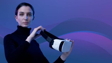 Elinde VR kulaklığı olan çekici genç bir kadın kameraya, sanal gerçekliğe ve yapay deneyim konseptine bakıyor.