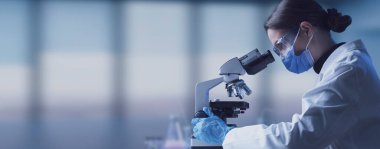 Cerrahi maske takan bir araştırmacı mikroskop, tıbbi araştırma ve bilim konseptine bakıyor.