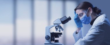 Laboratuvarda yüz maskesi takmış dalgın bir araştırmacı mikroskop kullanıyor: tıbbi araştırmaların zorlukları ve zorlukları.