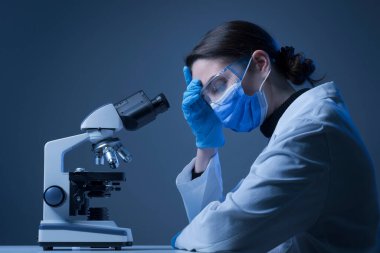 Laboratuvarda yüz maskesi takmış dalgın bir araştırmacı mikroskop kullanıyor: tıbbi araştırmaların zorlukları ve zorlukları.