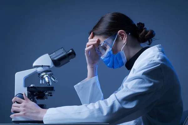 在实验室里戴着口罩的忧心忡忡的研究人员正在使用显微镜 医学研究的困难和挑战 — 图库照片