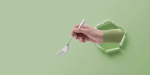Female hand holding a fork and coming out of a hole in paper, eating and restaurants concept