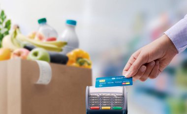 Bakkal parasını kredi kartıyla ödeyen bir kadın, kredi kartını POS terminalinin üzerinde tutuyor, arka planda alışveriş torbası var.