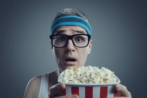 Funny guy with glasses and headband watching movies, he is staring at the screen with mouth open and eating popcorn